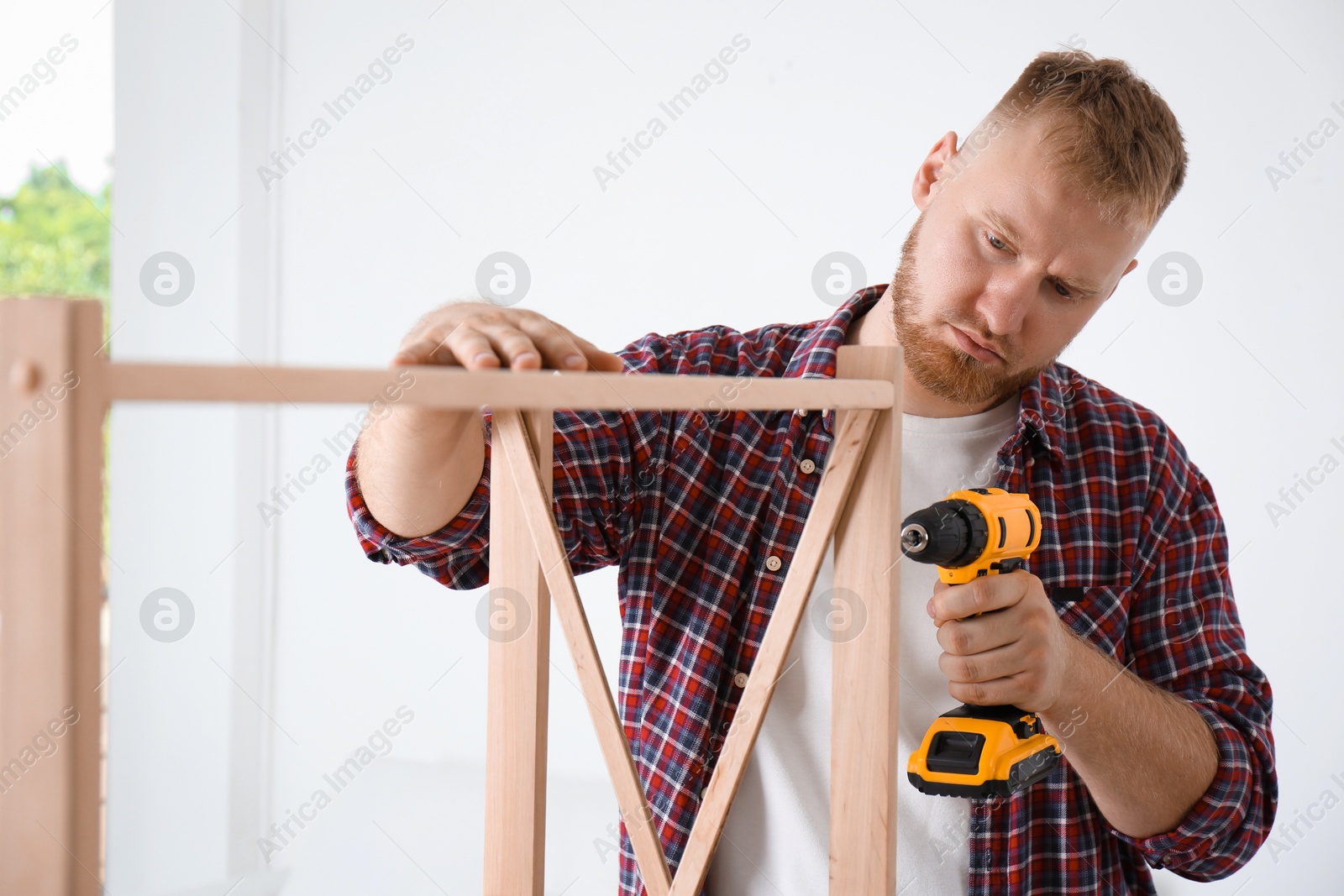 Photo of Man with electric screwdriver assembling wooden shelving unit at home