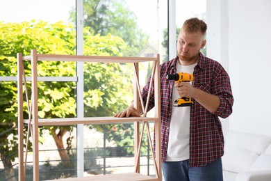 Photo of Man with electric screwdriver assembling wooden shelving unit at home