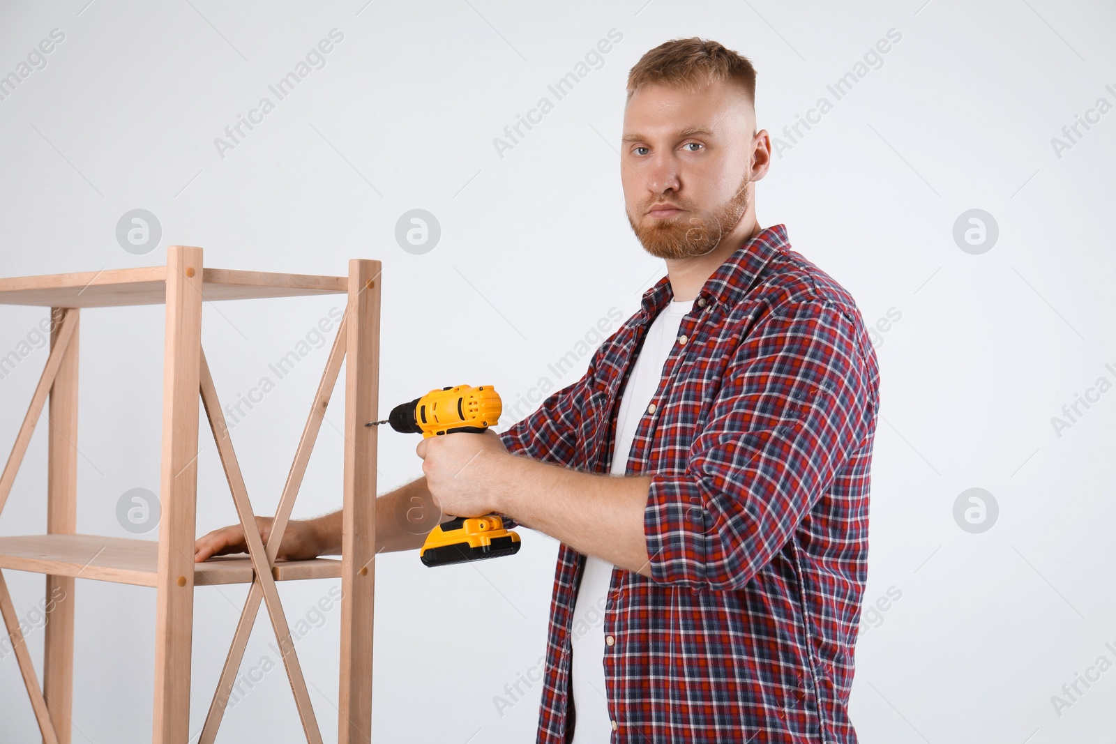 Photo of Man with electric screwdriver assembling furniture near white wall indoors