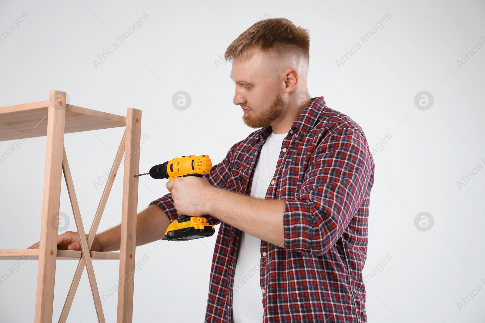 Photo of Man with electric screwdriver assembling furniture near white wall indoors