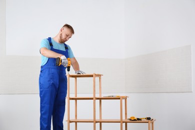 Worker with electric screwdriver assembling wooden shelving unit indoors