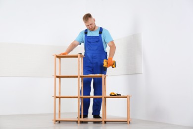 Photo of Worker with electric screwdriver assembling wooden shelving unit indoors