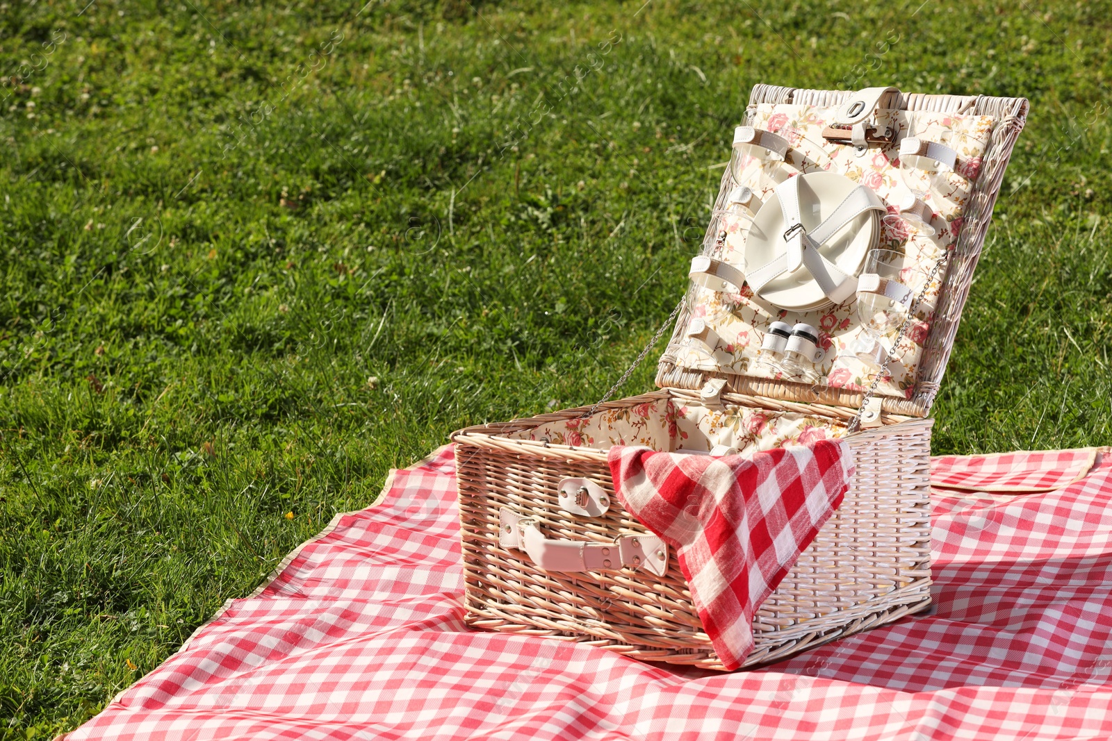 Photo of One picnic wicker basket with checkered napkin, tableware and blanket on green grass. Space for text