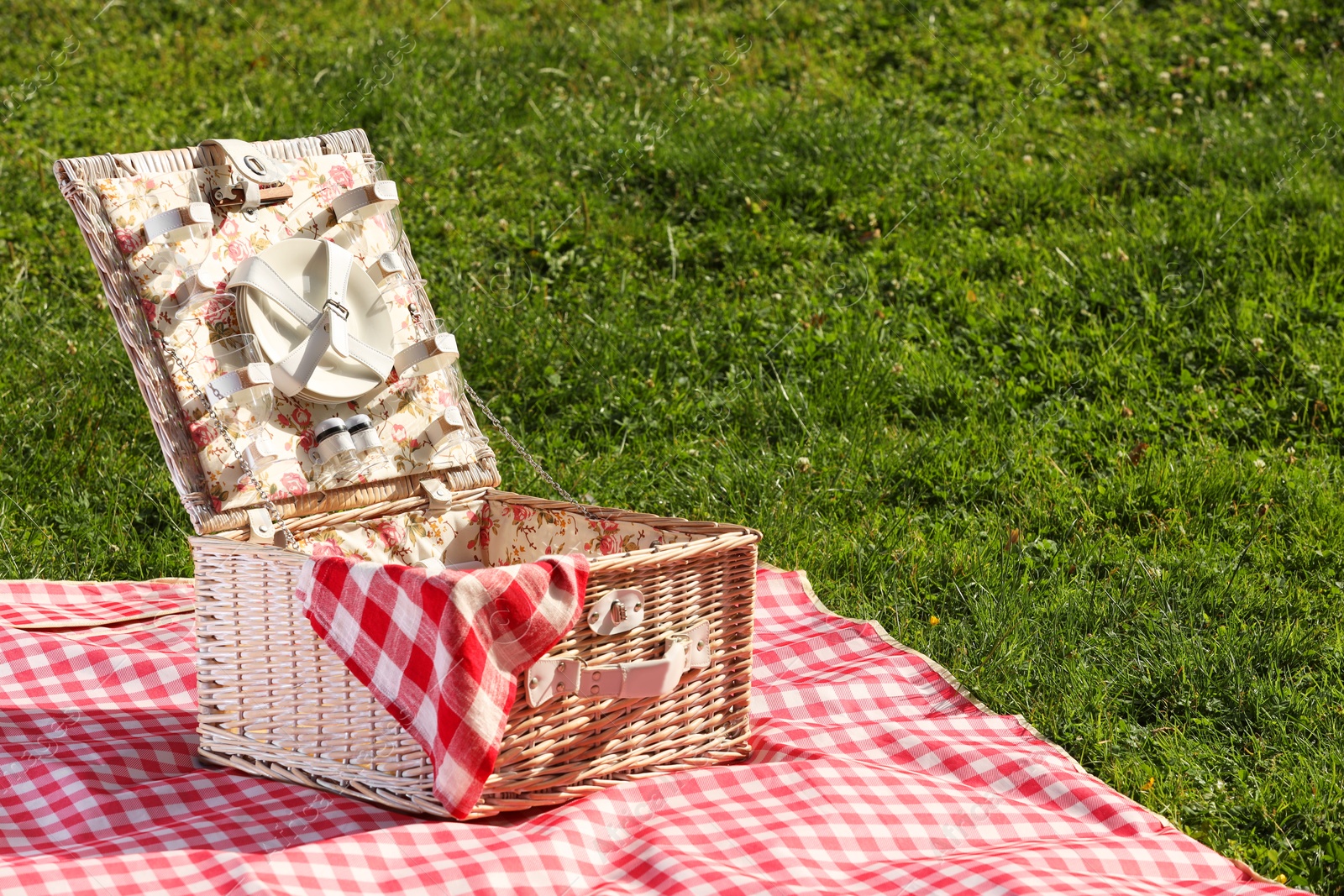 Photo of One picnic wicker basket with checkered napkin, tableware and blanket on green grass. Space for text