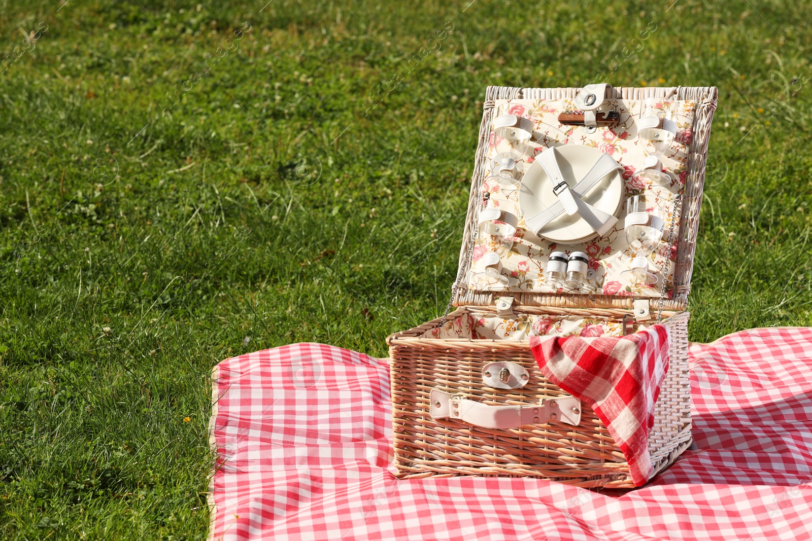 Photo of One picnic wicker basket with checkered napkin, tableware and blanket on green grass. Space for text