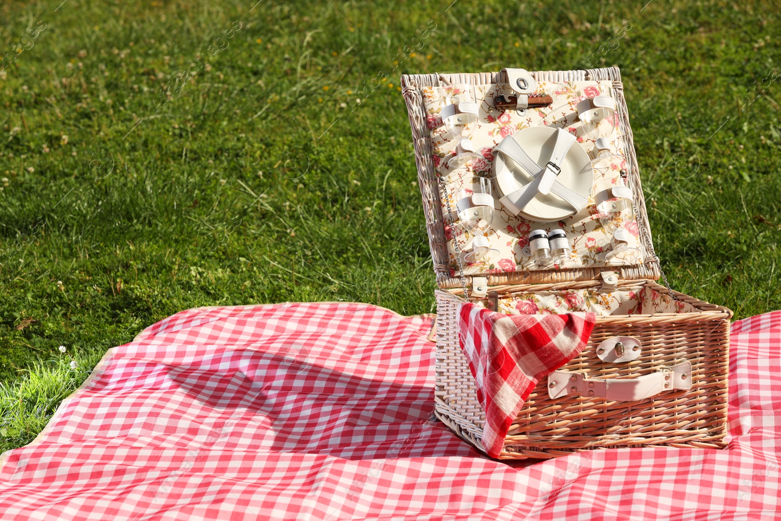 Photo of One picnic wicker basket with checkered napkin, tableware and blanket on green grass. Space for text