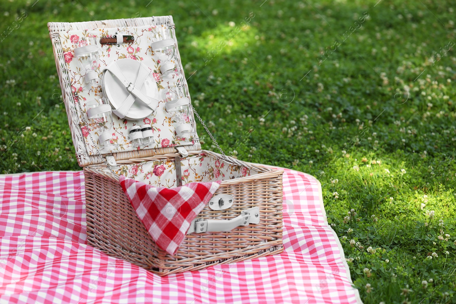 Photo of One picnic wicker basket with checkered napkin, tableware and blanket on green grass. Space for text