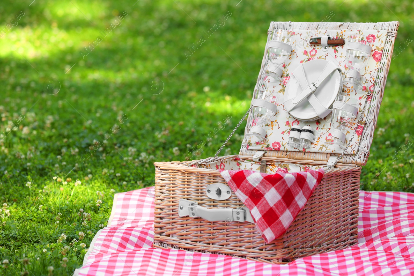 Photo of One picnic wicker basket with checkered napkin, tableware and blanket on green grass. Space for text