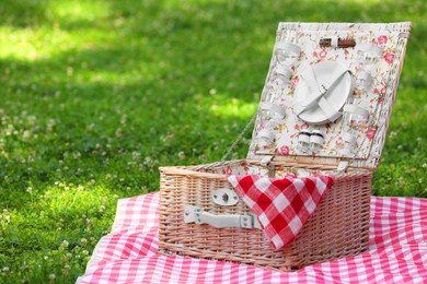 Photo of One picnic wicker basket with checkered napkin, tableware and blanket on green grass. Space for text