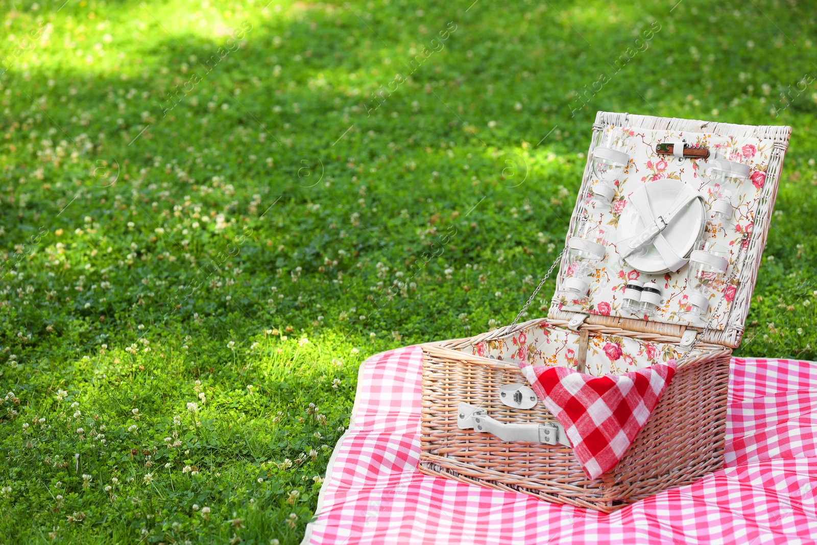 Photo of One picnic wicker basket with checkered napkin, tableware and blanket on green grass. Space for text