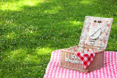 Photo of One picnic wicker basket with checkered napkin, tableware and blanket on green grass. Space for text