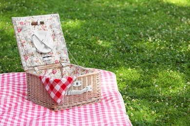 Photo of One picnic wicker basket with checkered napkin, tableware and blanket on green grass. Space for text