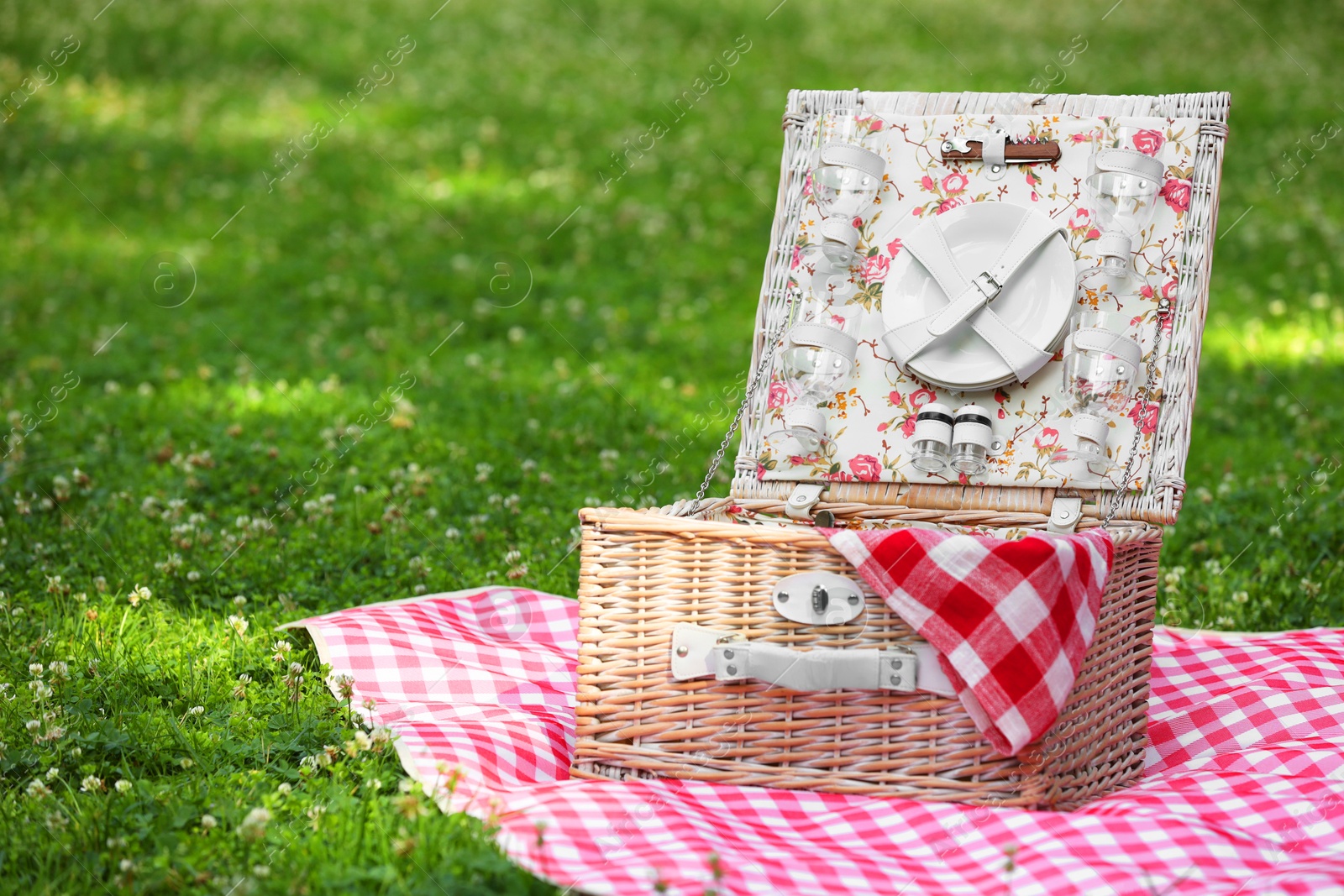 Photo of One picnic wicker basket with checkered napkin, tableware and blanket on green grass. Space for text