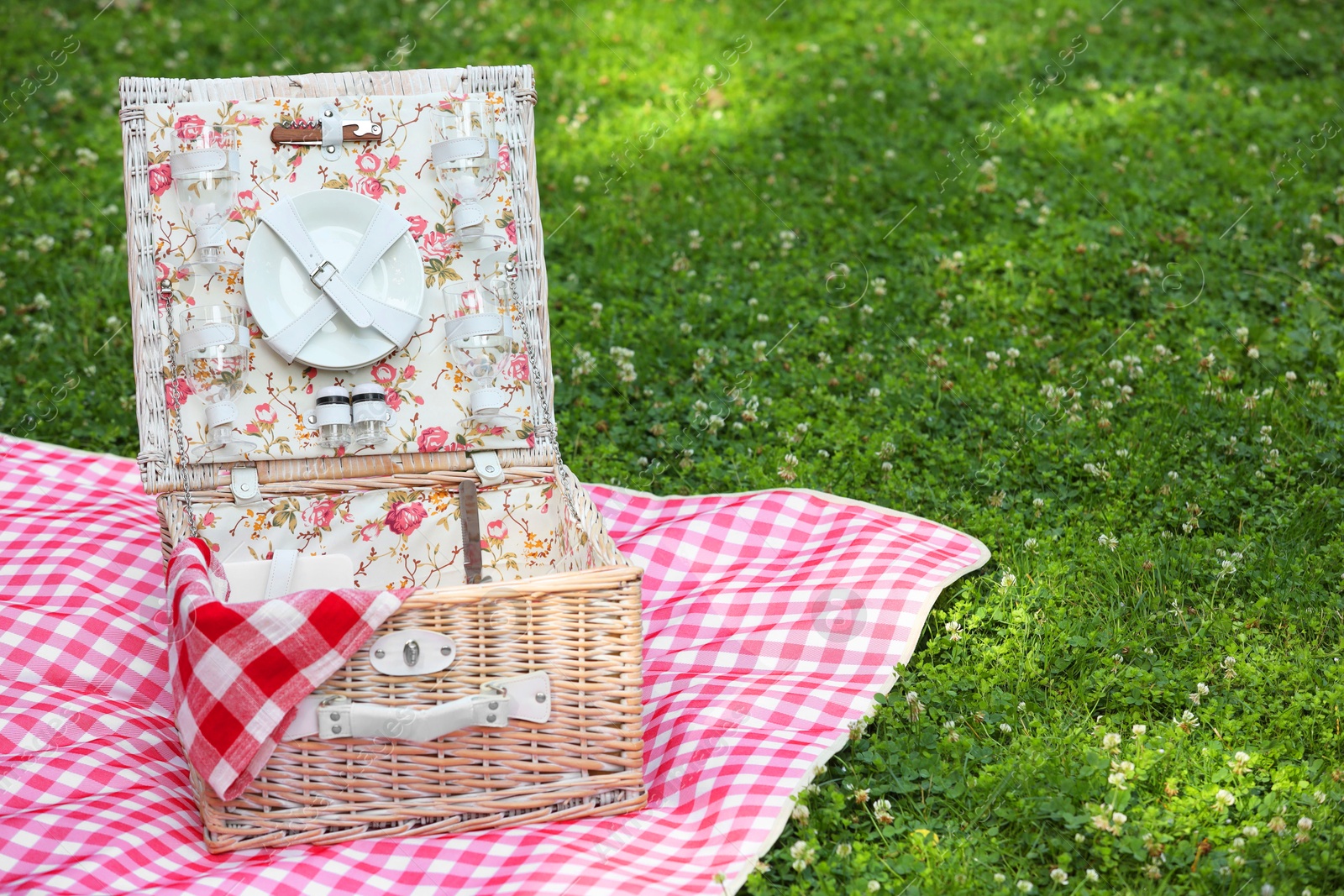 Photo of One picnic wicker basket with checkered napkin, tableware and blanket on green grass. Space for text