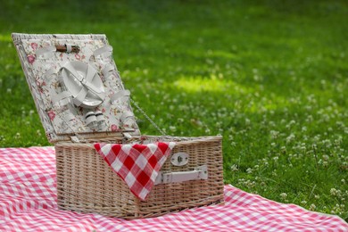 Photo of One picnic wicker basket with checkered napkin, tableware and blanket on green grass. Space for text