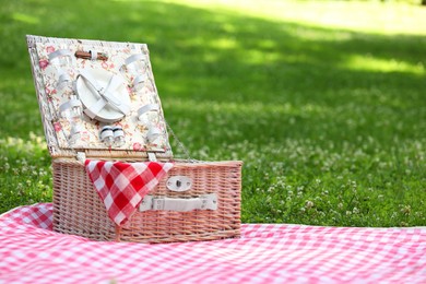 Photo of One picnic wicker basket with checkered napkin, tableware and blanket on green grass. Space for text