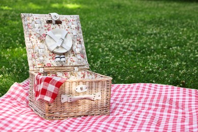 Photo of One picnic wicker basket with checkered napkin, tableware and blanket on green grass. Space for text