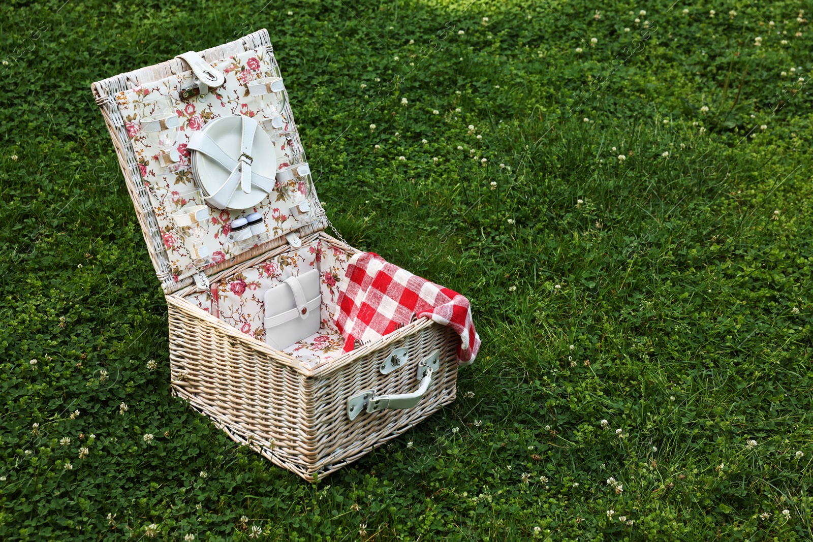 Photo of One picnic wicker basket with checkered napkin and tableware on green grass. Space for text