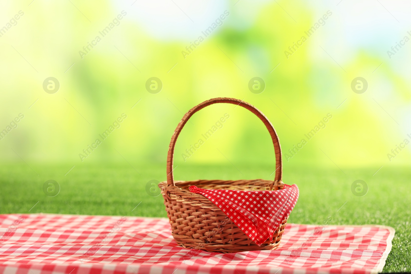 Photo of One picnic wicker basket with checkered napkin and blanket on grass against blurred green background. Space for text