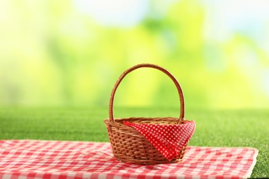 Photo of One picnic wicker basket with checkered napkin and blanket on grass against blurred green background. Space for text