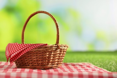 Photo of One picnic wicker basket with checkered napkin and blanket on grass against blurred green background, closeup. Space for text