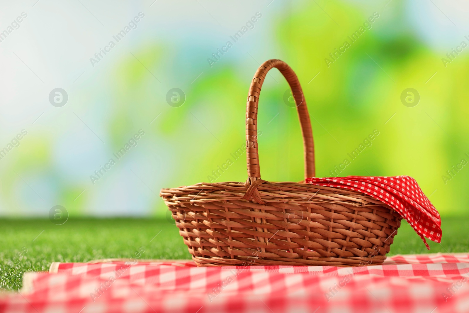 Photo of One picnic wicker basket with checkered napkin and blanket on grass against blurred green background. Space for text