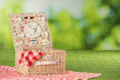 Photo of One picnic wicker basket with checkered napkin, tableware and blanket on grass against blurred green background. Space for text