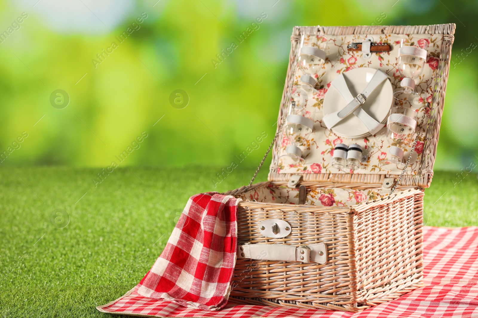 Photo of Picnic wicker basket with checkered napkin, tableware and blanket on grass against blurred green background. Space for text