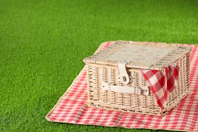 Photo of One picnic wicker basket with checkered napkin and blanket on green grass, closeup. Space for text