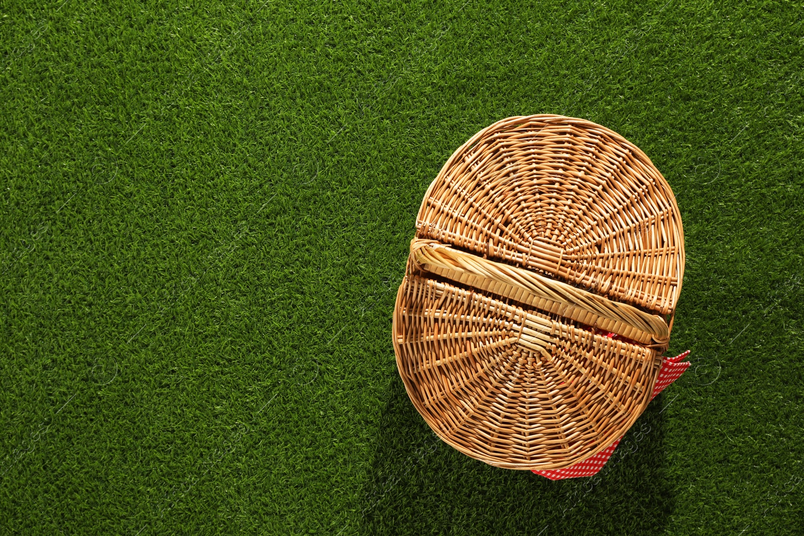 Photo of One picnic wicker basket with checkered napkin on green grass, top view. Space for text