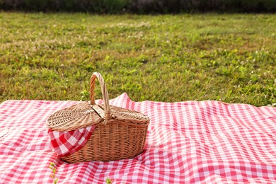 Photo of Picnic wicker basket with napkin and red checkered blanket on green grass outdoors, space for text