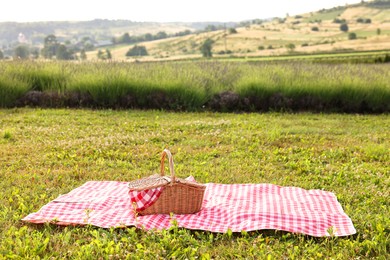 Photo of Picnic wicker basket with napkin and red checkered blanket on green grass outdoors