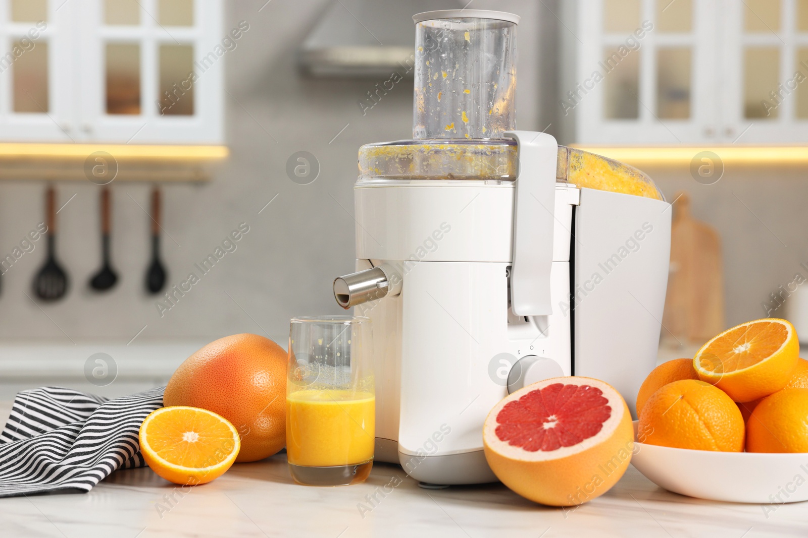 Photo of Modern juicer, oranges, grapefruits and glass on white marble table in kitchen