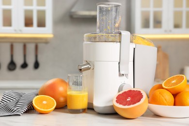 Modern juicer, oranges, grapefruits and glass on white marble table in kitchen