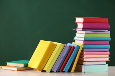 Photo of Stacks of many colorful books on white table near chalkboard in classroom. Space for text