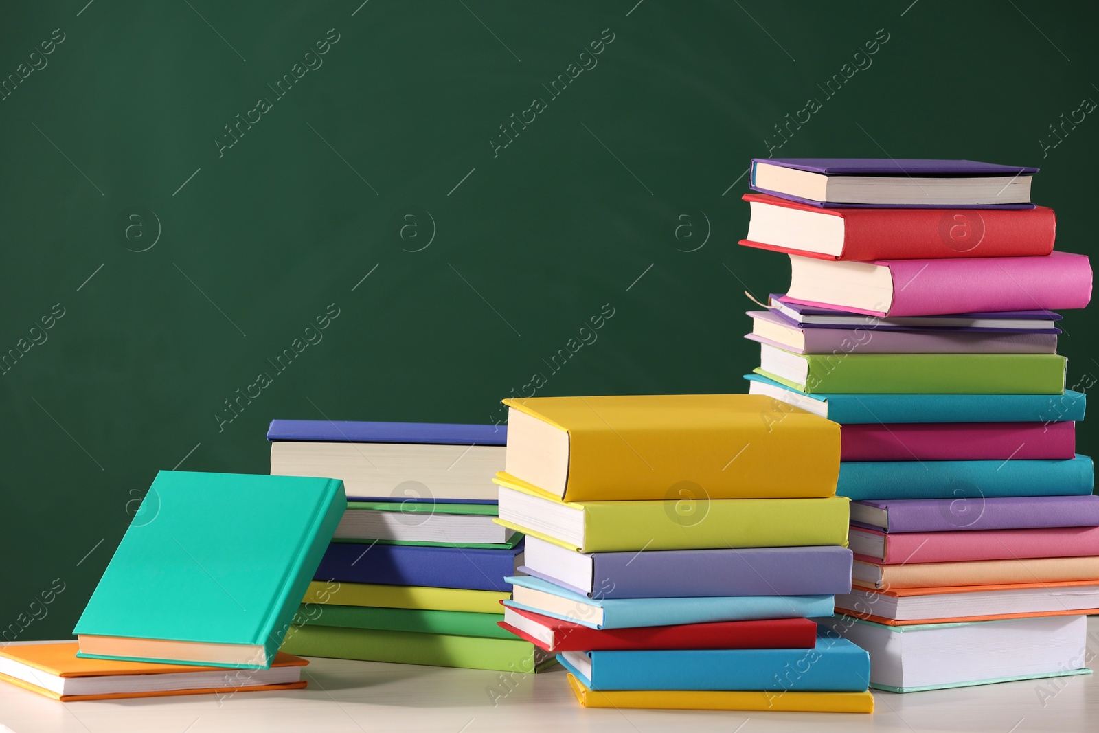 Photo of Stacks of many colorful books on white table near chalkboard in classroom. Space for text