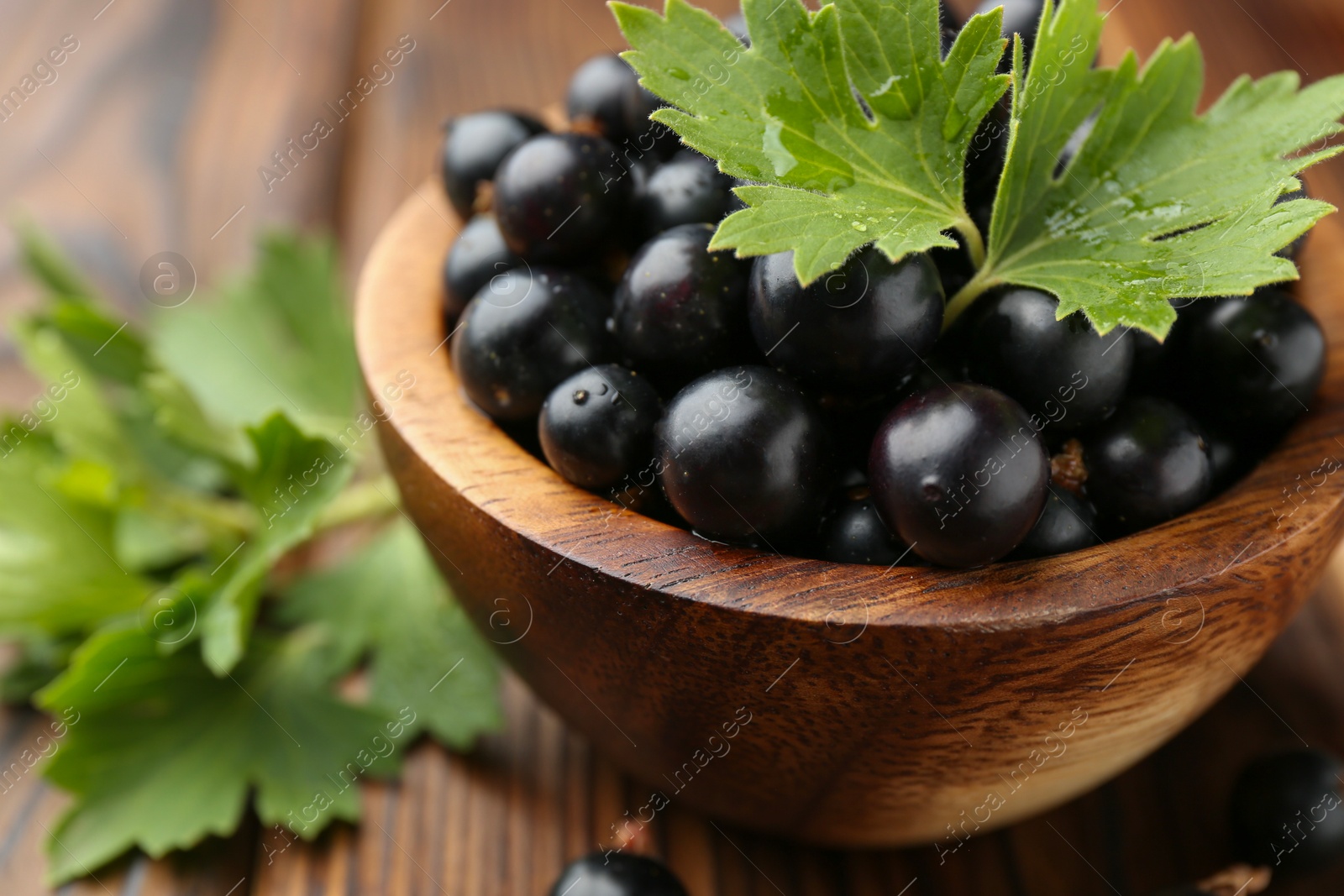 Photo of Ripe black currants and leaves in bowl on wooden table, closeup