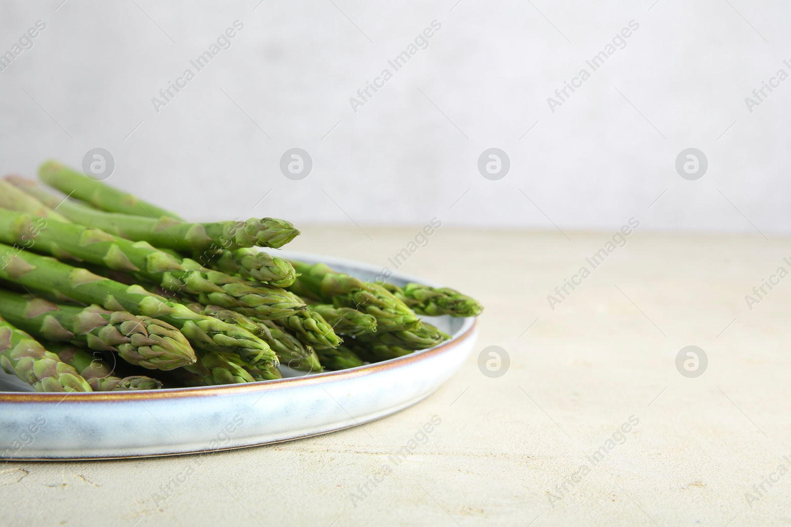 Photo of Plate with fresh green asparagus stems on light textured table, closeup. Space for text