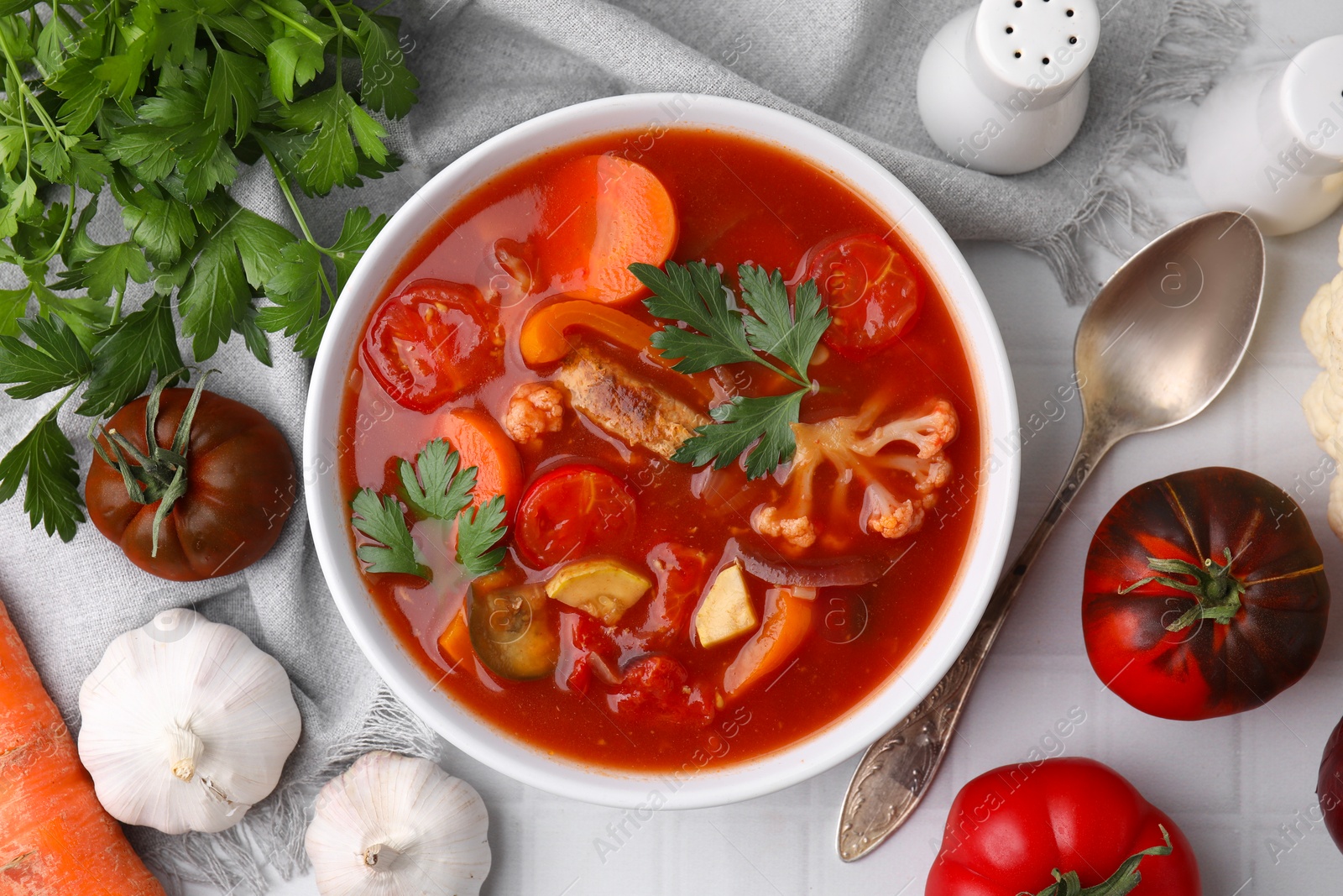 Photo of Delicious homemade stew in bowl, spoon and ingredients on white tiled table, flat lay