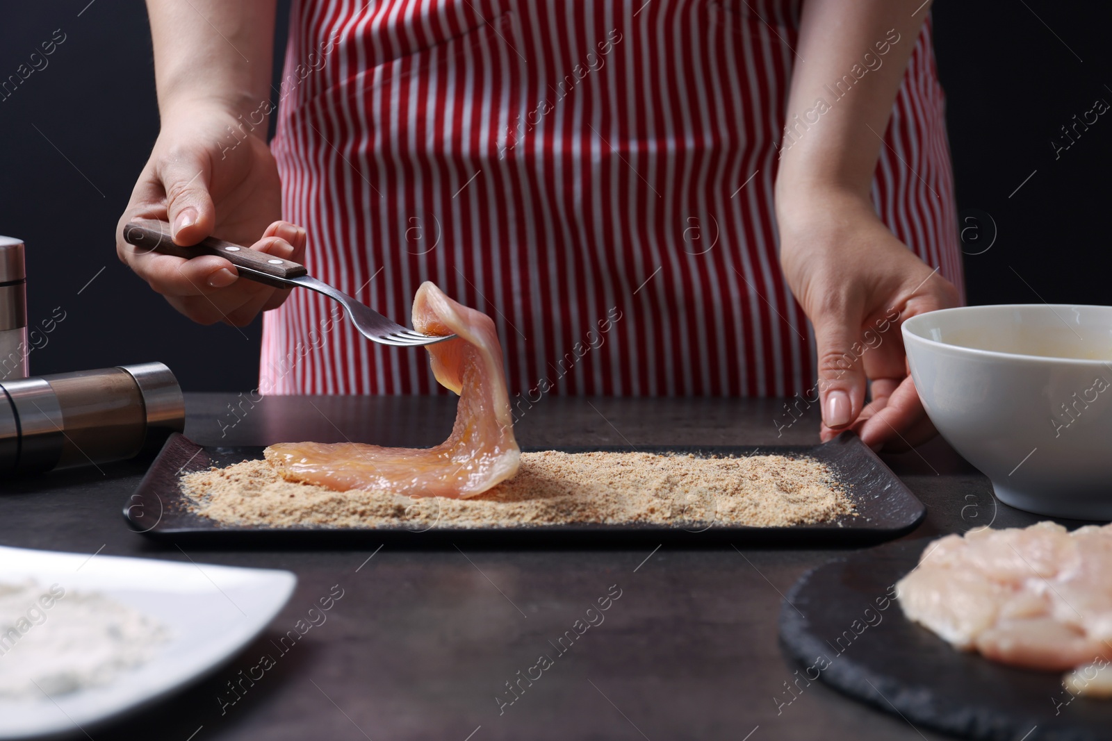 Photo of Making schnitzel. Woman coating slice of meat with bread crumbs at dark table, closeup