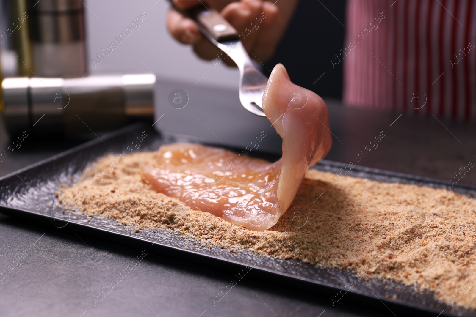 Photo of Making schnitzel. Woman coating slice of meat with bread crumbs at dark table, closeup