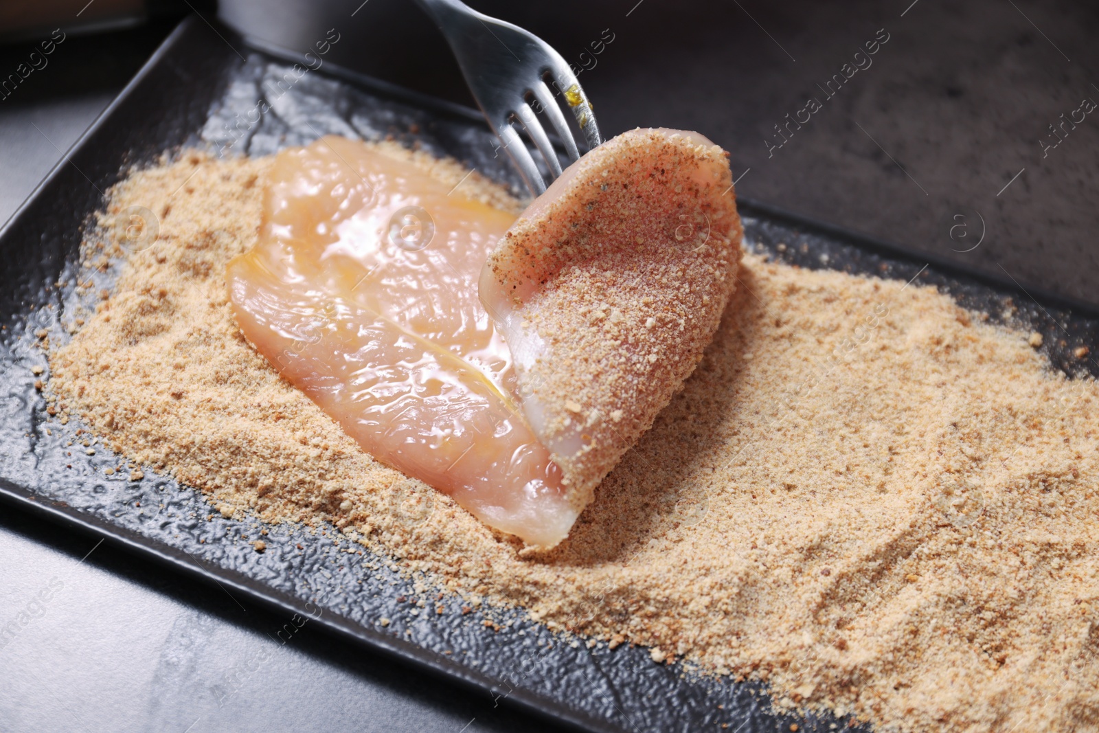 Photo of Making schnitzel. Coating slice of meat with bread crumbs at dark table, closeup