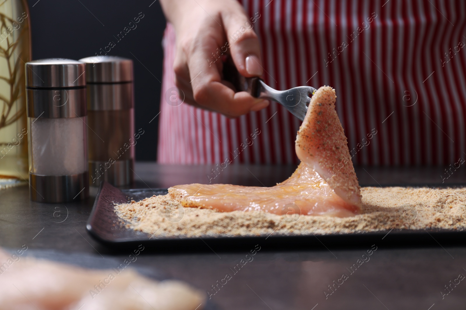 Photo of Making schnitzel. Woman coating slice of meat with bread crumbs at dark table, closeup