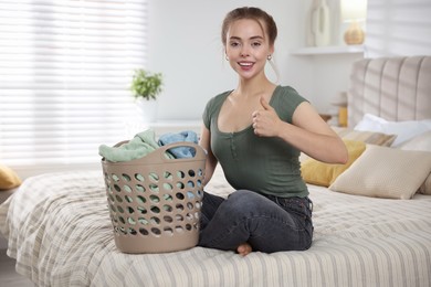 Photo of Happy young housewife with basket full of laundry showing thumbs up on bed at home