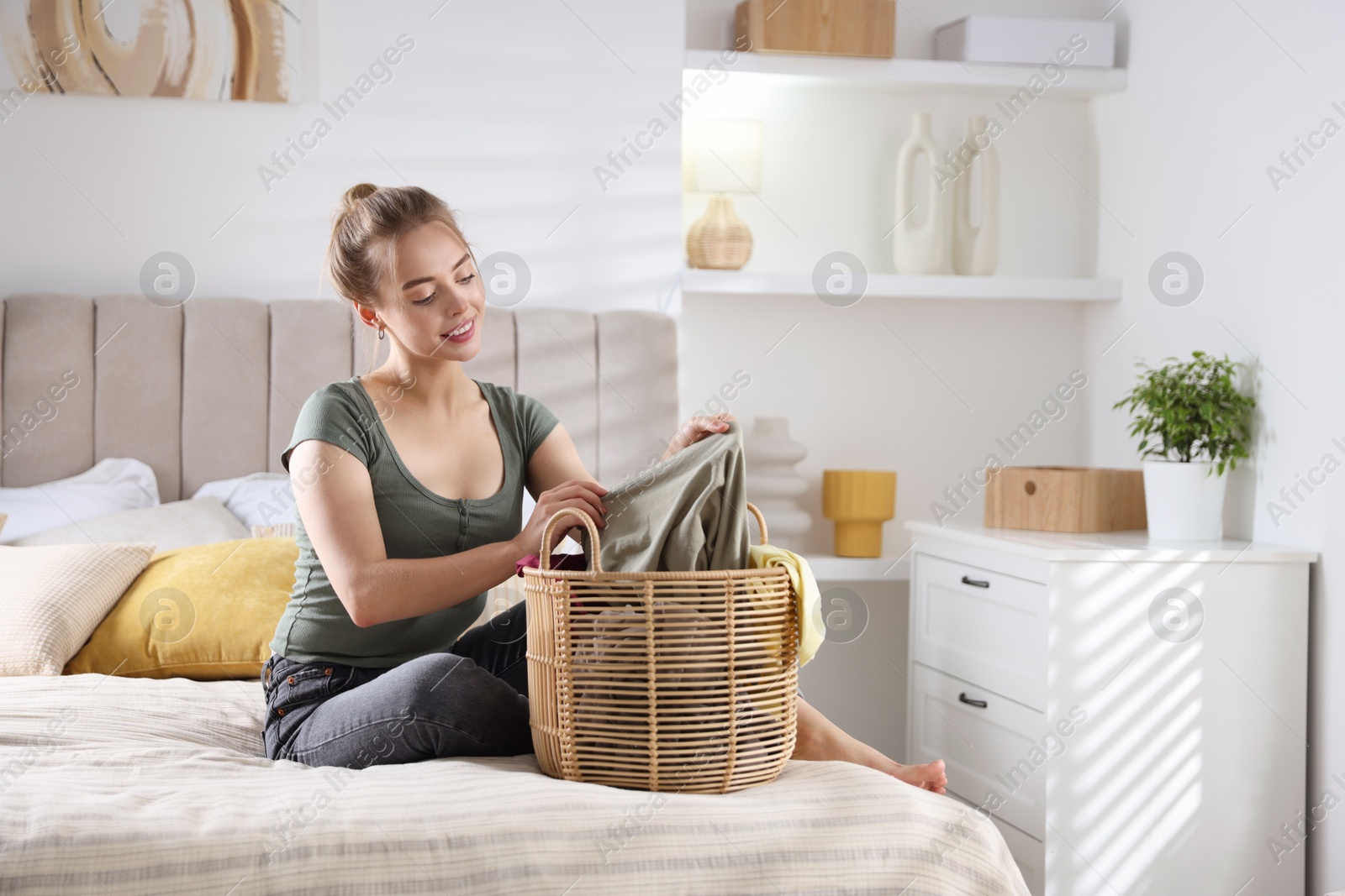 Photo of Happy young housewife with basket full of laundry on bed at home