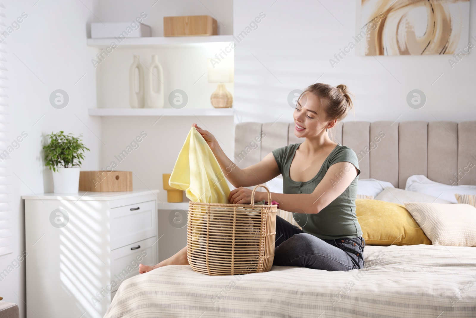 Photo of Happy young housewife with basket full of laundry on bed at home