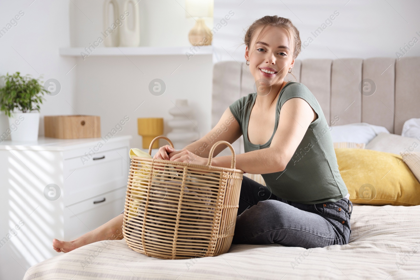 Photo of Happy young housewife with basket full of laundry on bed at home