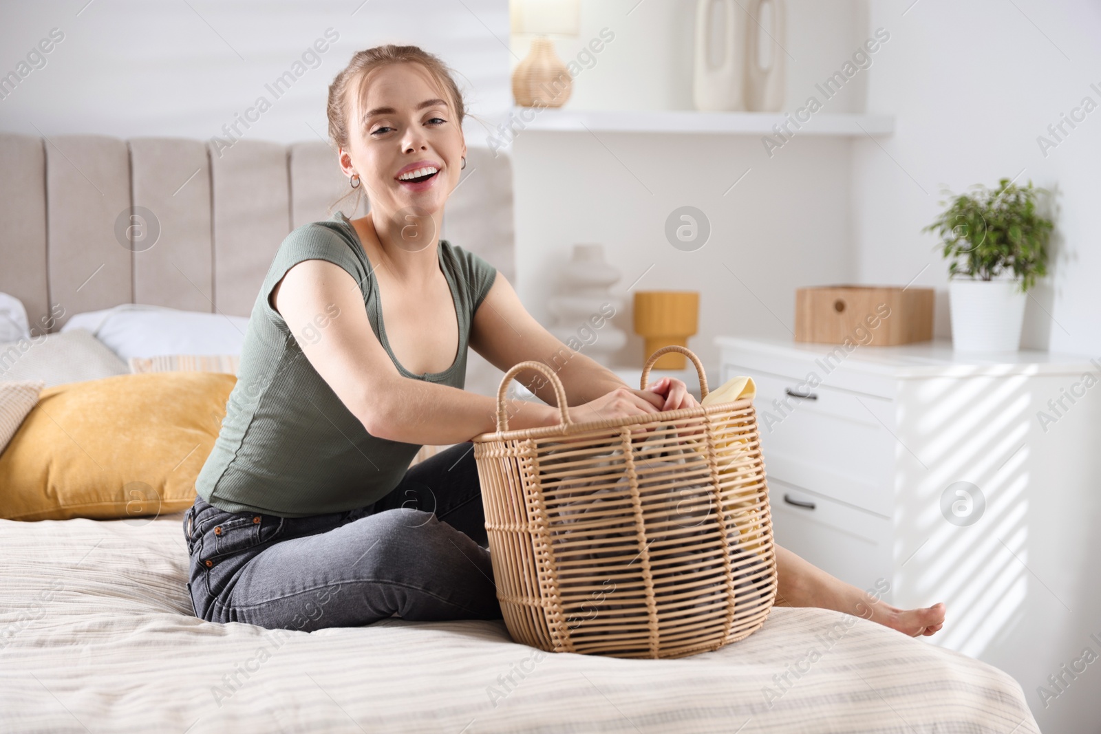 Photo of Happy young housewife with basket full of laundry on bed at home