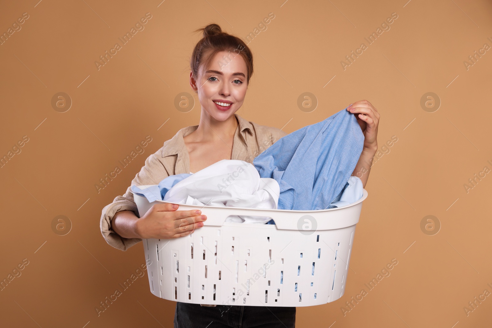 Photo of Happy young housewife with basket full of laundry on pale orange background