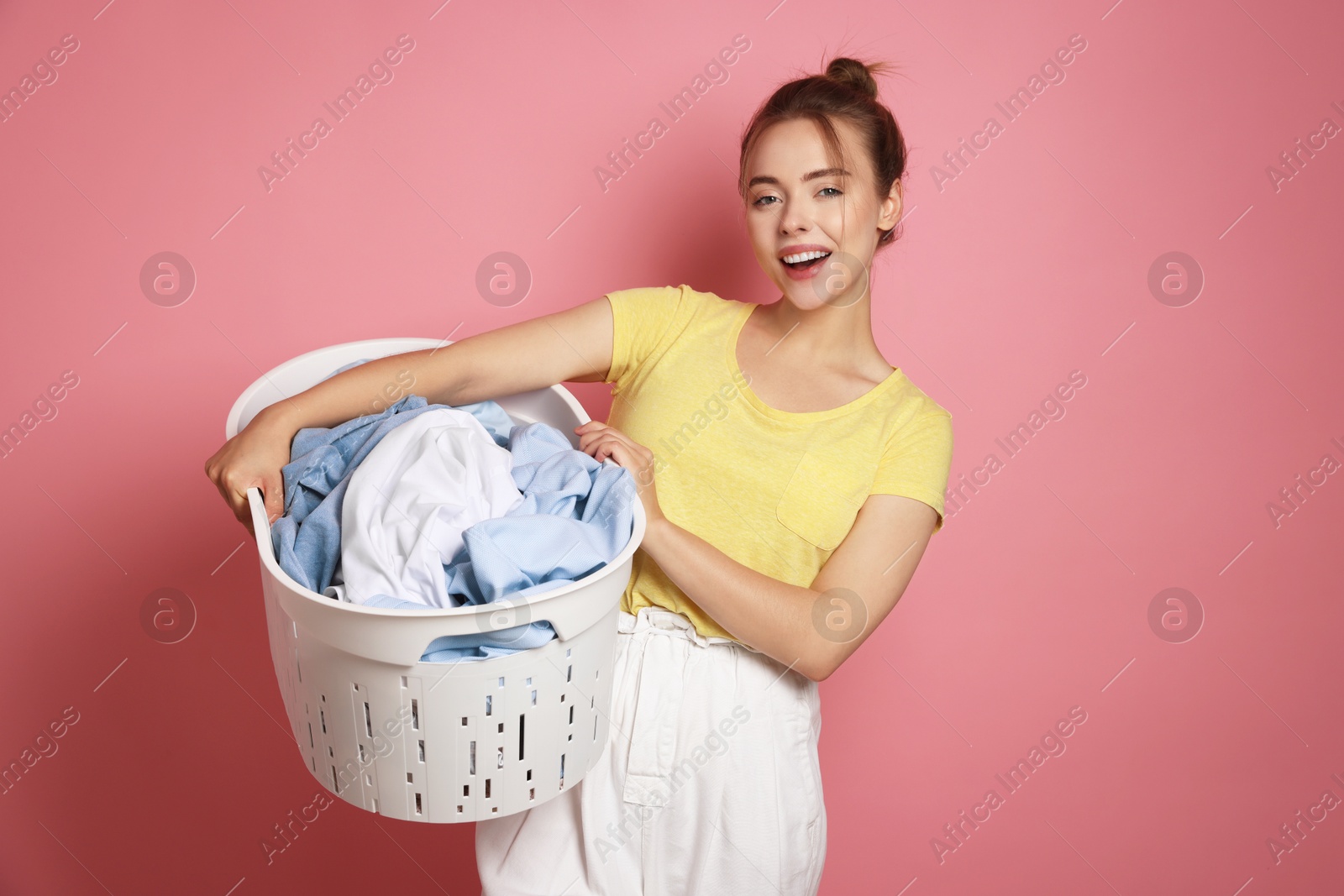 Photo of Happy young housewife with basket full of laundry on pale pink background